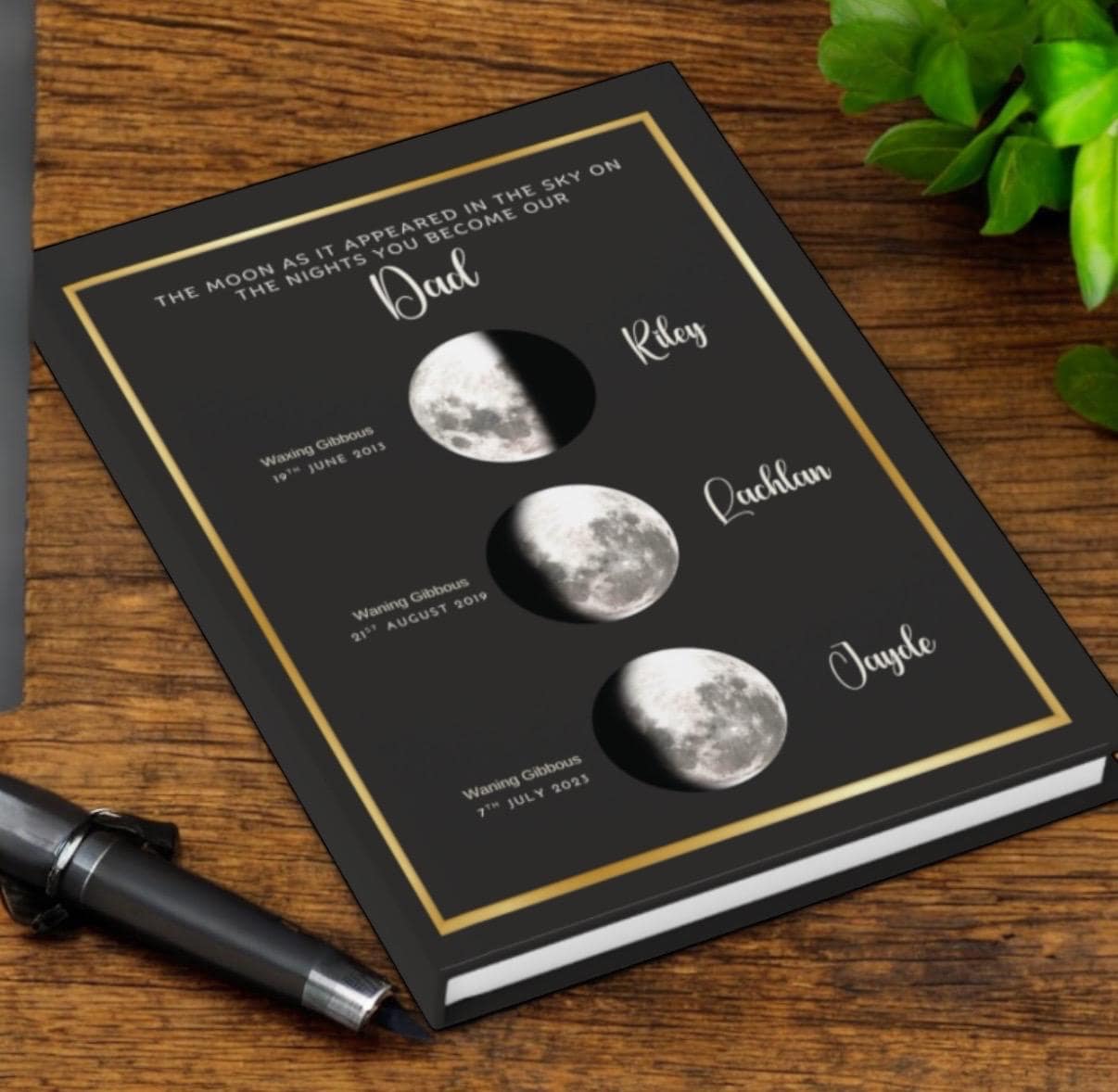 A personalized notebook with moon phases, names (Dad, Riley, Lachlan, Jayde), and dates on the cover, placed on a wooden desk next to a pen and a green plant.