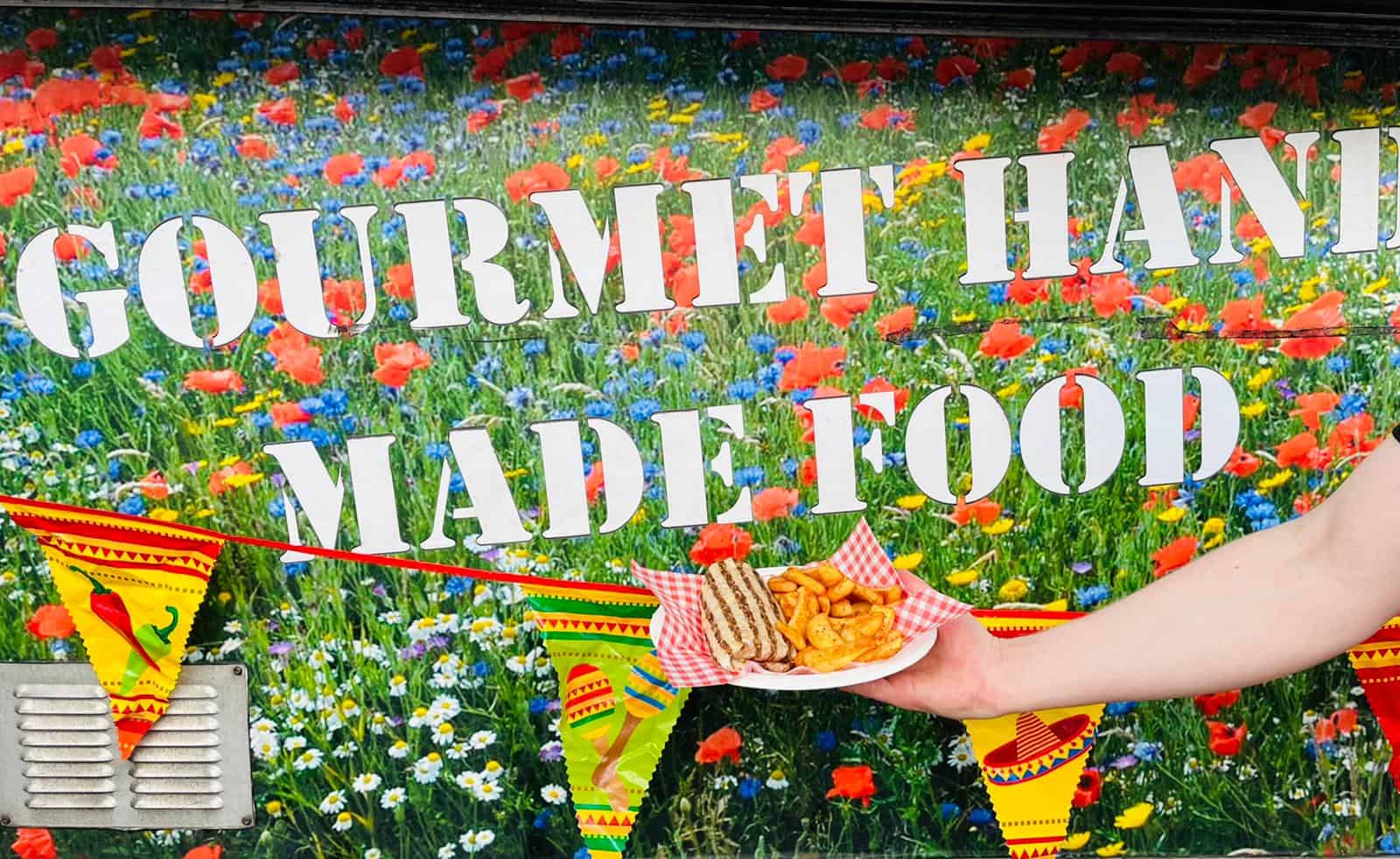 Colourful field of wildflowers with "GOUÎMET HALL MADE FOOD" sign promoting catering services, vibrant and inviting.
