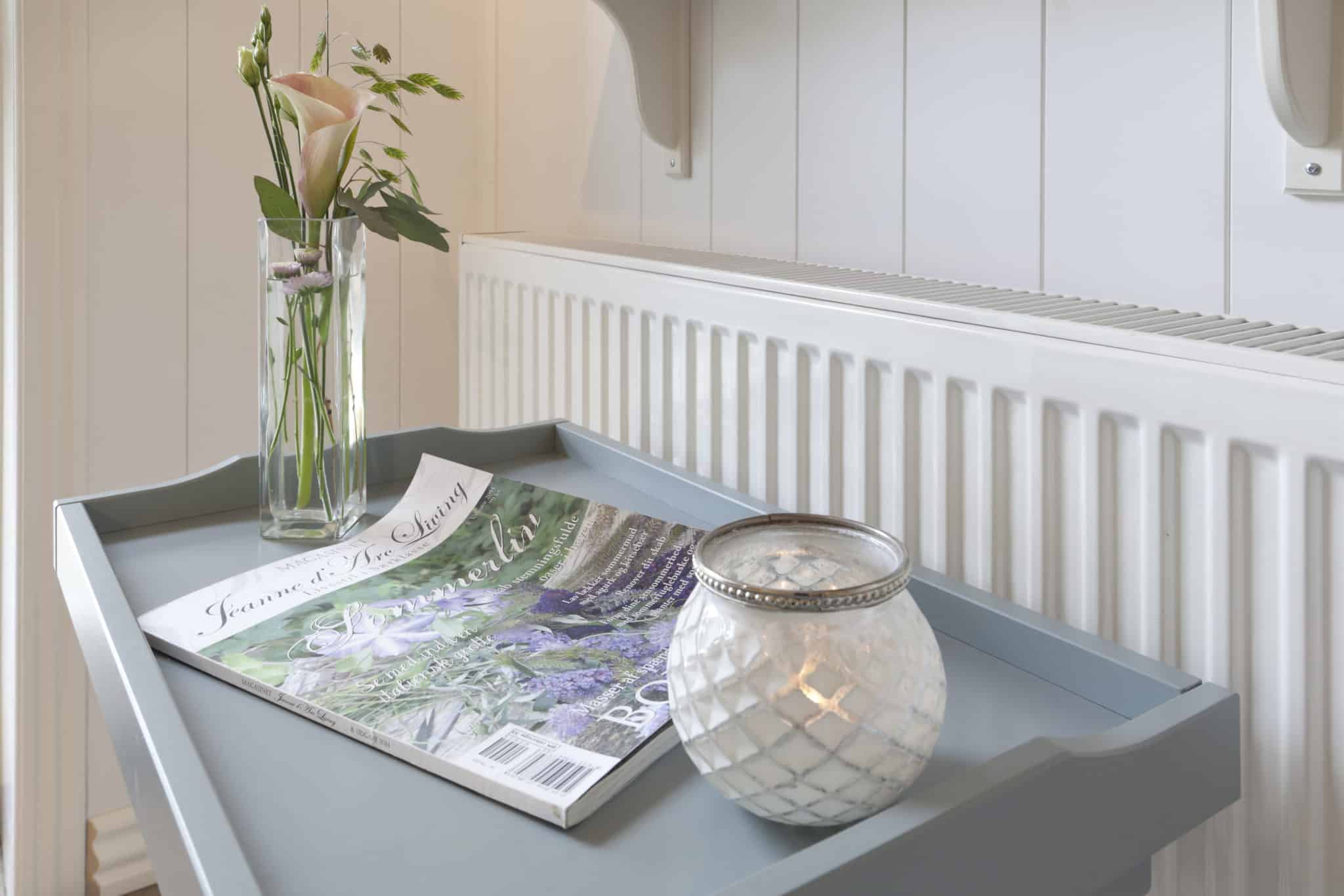 Hvide vægpaneler og radiator, vase med blomster, lampe og bladre til afslapning på Badehotel.