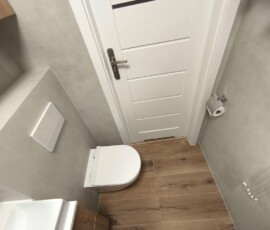 Aranżacja typowej małej łazienki w bloku na Gocławiu. Styl natura – płytki drewnopodobne, jasny gres, czarna armatura i prysznic walk-in.