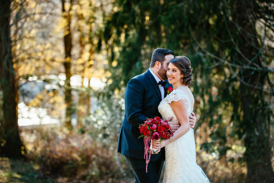 Bride and groom embracing in a wooded setting with a vibrant wedding bouquet.