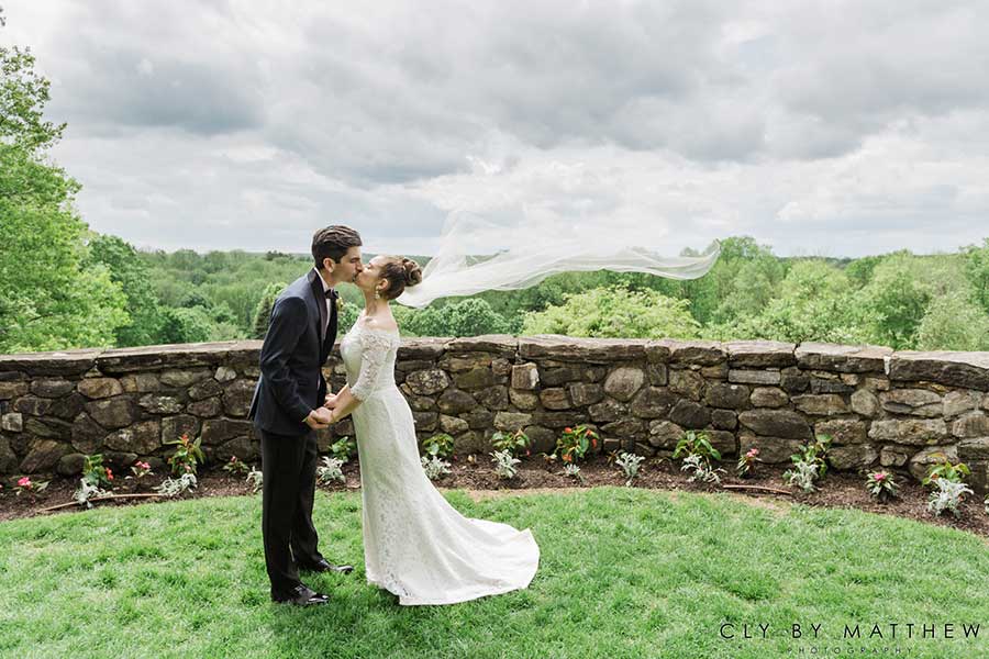 Bride and groom sharing a kiss with flowing veil and scenic outdoor venue backdrop.
