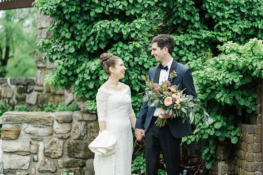 Bride and groom holding hands beside lush greenery at an outdoor wedding venue.