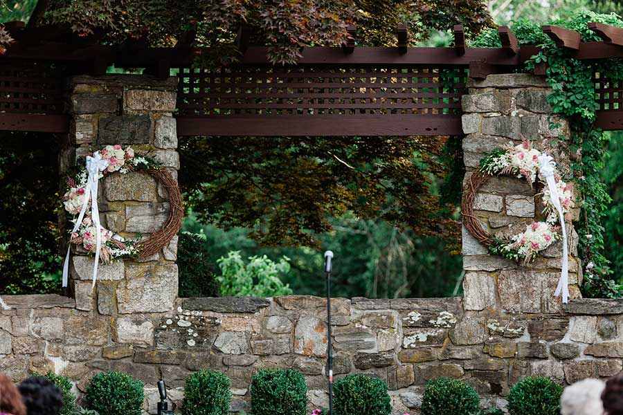Outdoor wedding ceremony backdrop with floral wreaths and stone garden pergola.