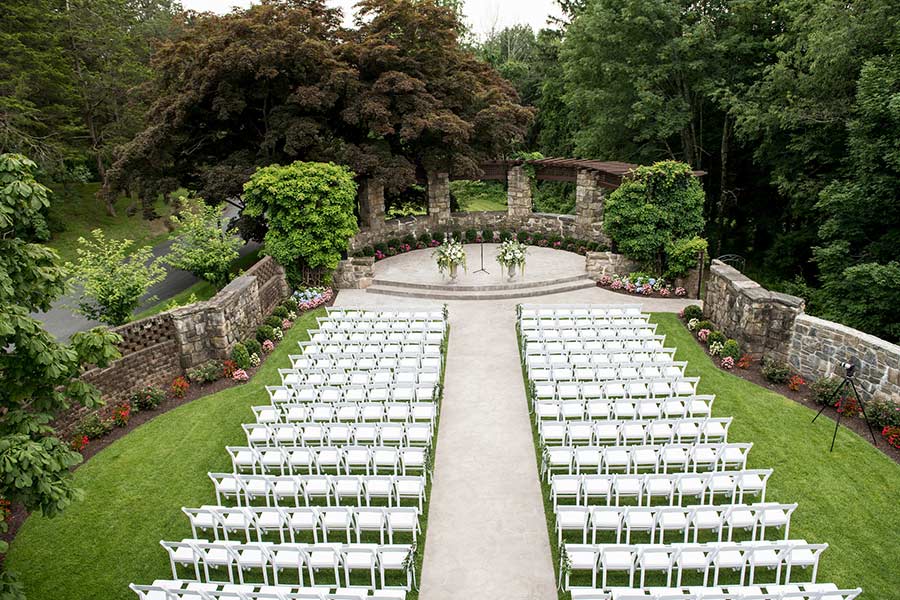 Outdoor wedding ceremony space with garden seating and a stone pergola backdrop.
