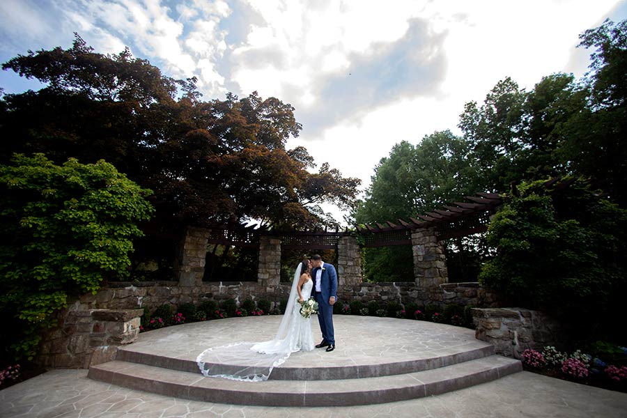 A bride and groom sharing a kiss on an outdoor stone patio venue, framed by stone pillars, a wooden pergola, and lush green trees under a partly cloudy sky.