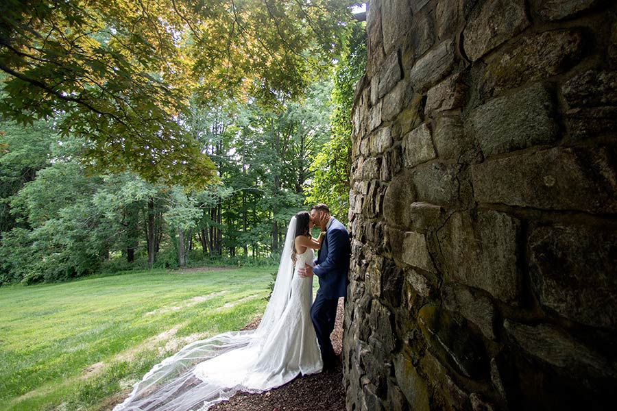 Bride and groom sharing an intimate moment beside a rustic stone wall and garden lawn.