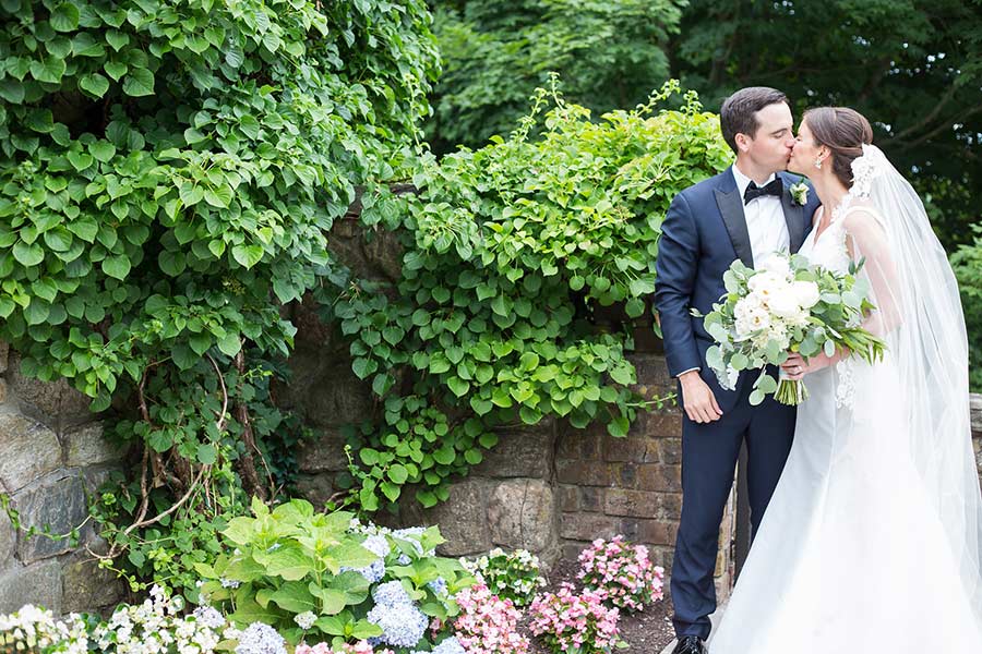 Bride and groom kissing beside lush garden greenery at a romantic wedding venue.