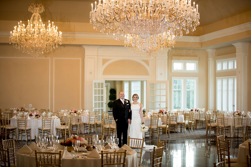 Bride and groom standing in an elegant ballroom beneath grand crystal chandeliers.