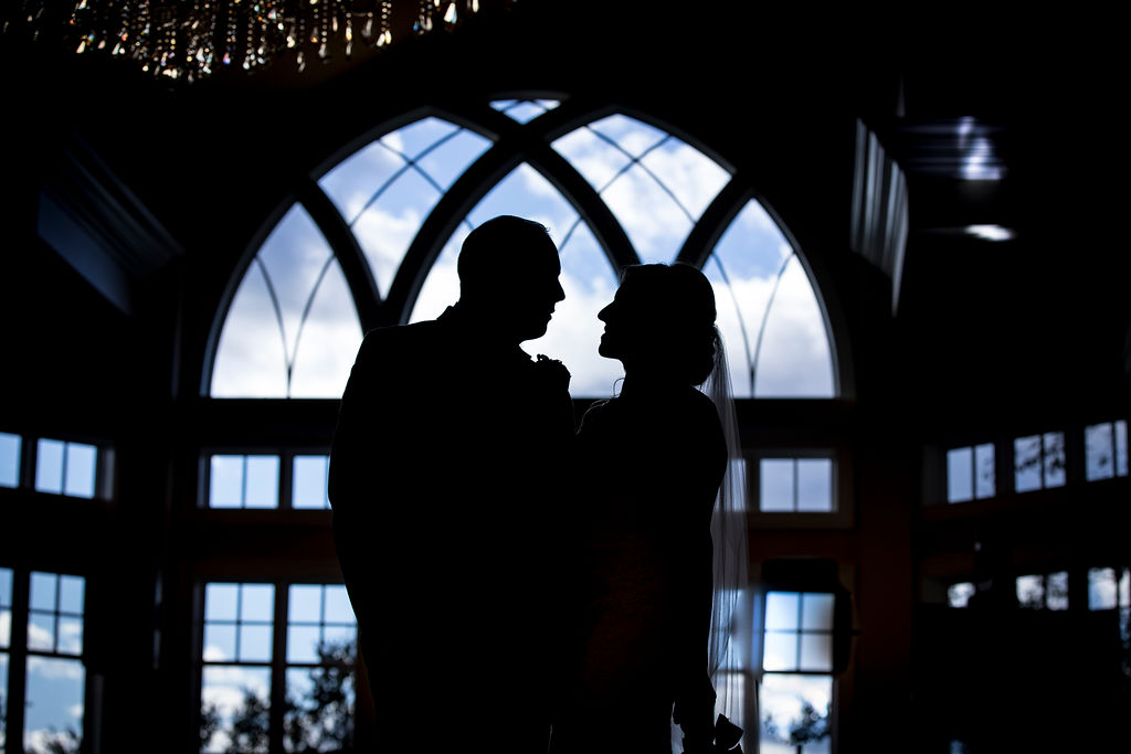 Bride and groom silhouette beneath grand windows in an elegant wedding venue.