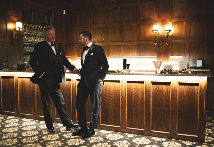 Grooms standing by elegant wood bar at Le Chateau wedding reception venue