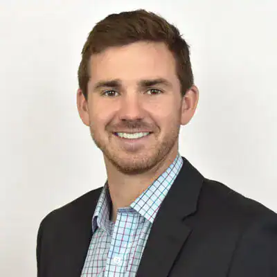 Headshot of a smiling young man with brown hair, wearing a dark blazer and a checkered shirt, against a plain white background.