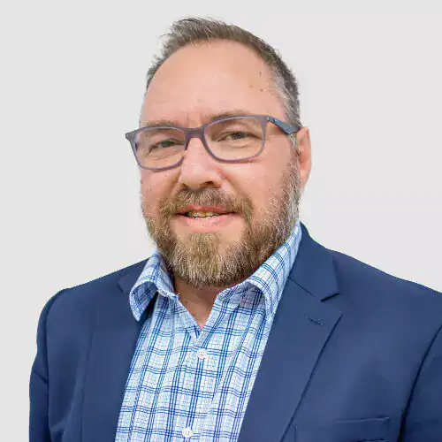 Close-up of a middle-aged man with glasses, beard, and short hair, wearing a blue suit and checkered shirt, smiling at the camera.