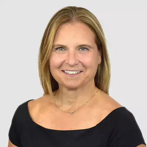 A professional woman with blonde hair smiling at the camera, wearing a black top and a delicate necklace, against a plain light background.