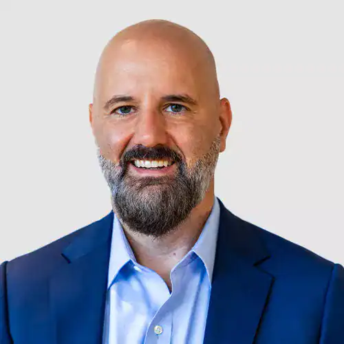 A professional businessman with a beard and blue eyes, wearing a navy suit and light blue shirt, smiling at the camera against a plain light background.