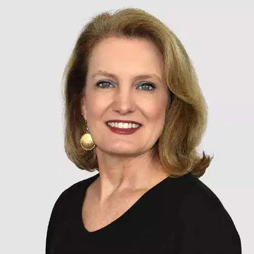 A professional woman with blonde hair, wearing a black top and gold earrings, smiling at the camera against a plain light background.