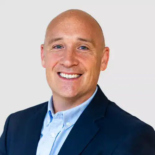 Close-up of a smiling man with a bald head, wearing a navy blazer and light blue shirt, against a plain light background.