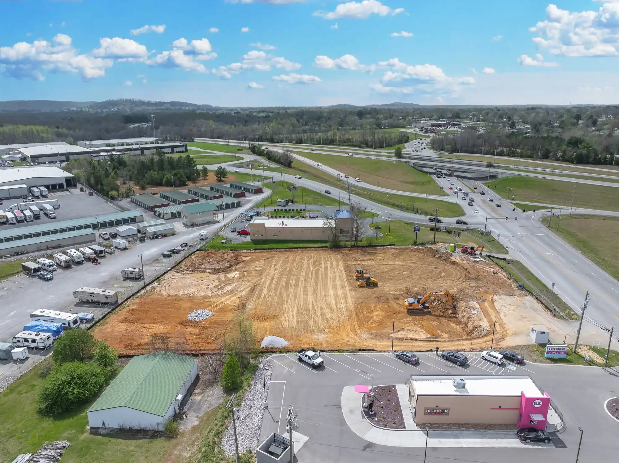 An aerial view of a construction site with heavy machinery and cleared land near a busy highway and commercial buildings.