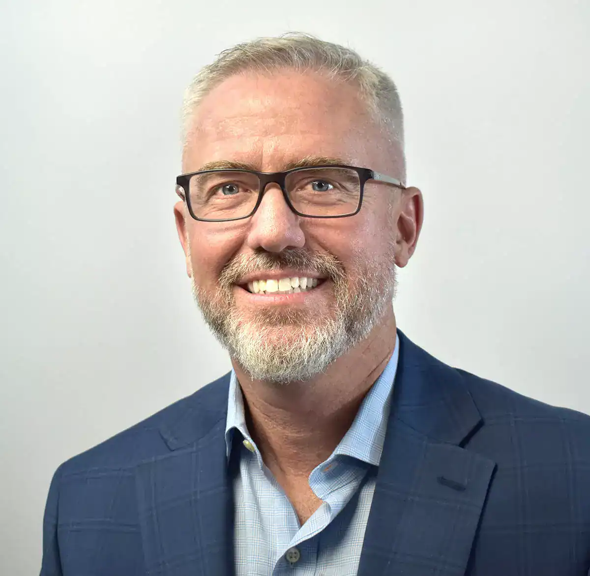 Close-up of a smiling middle-aged man with glasses, gray hair, and a beard, wearing a blue suit and light blue shirt against a plain background.