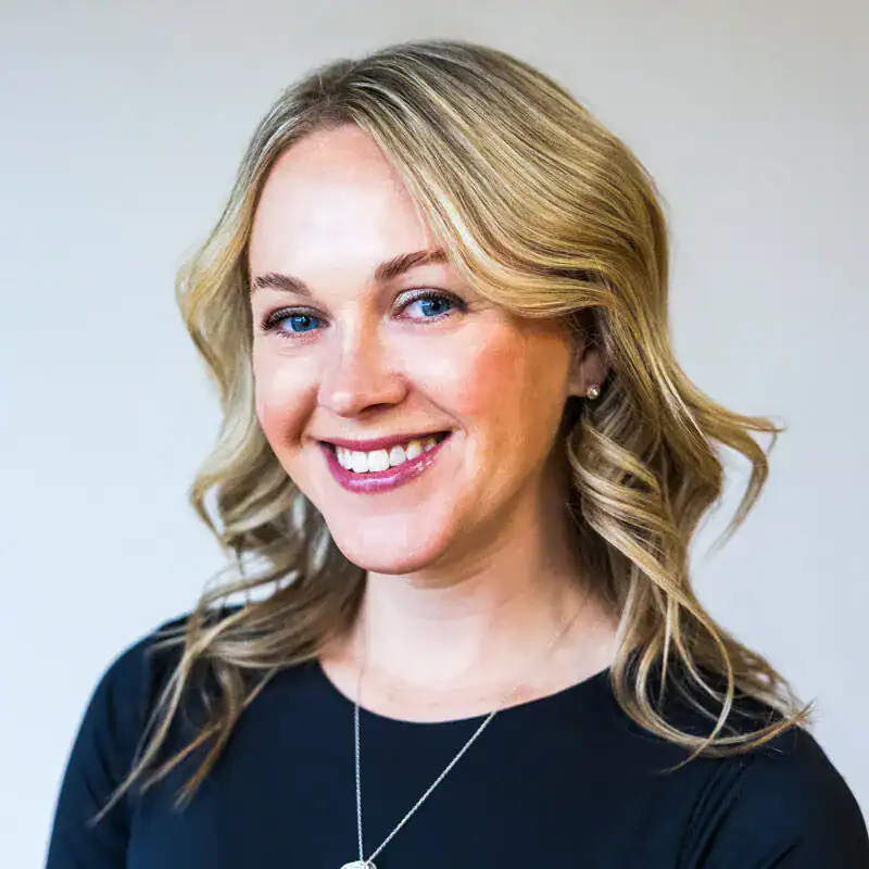 A smiling woman with blonde wavy hair wearing a black top and a silver necklace, looking directly at the camera against a plain background.