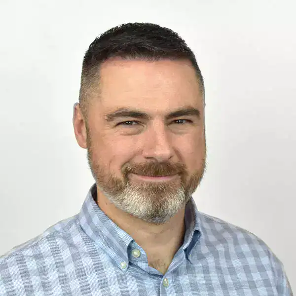 Close-up portrait of a man with short dark hair, a beard, and a friendly smile, wearing a light blue checkered shirt against a plain white background.