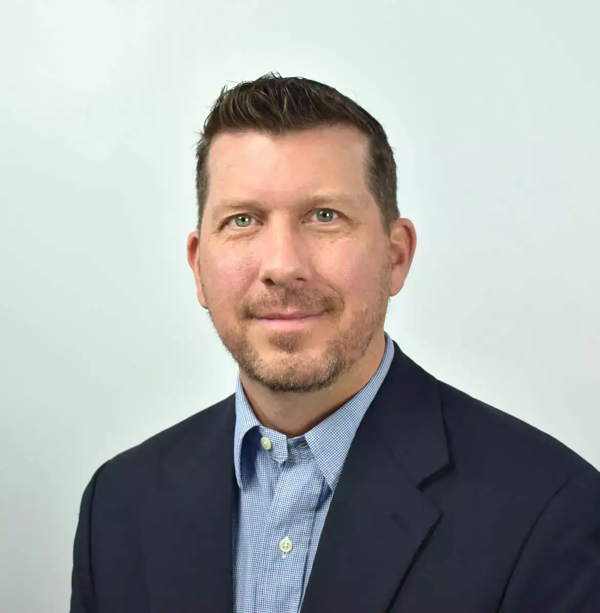 A professional businessman with short dark hair, blue eyes, and a light beard, wearing a navy blazer and light blue shirt, smiling slightly at the camera.