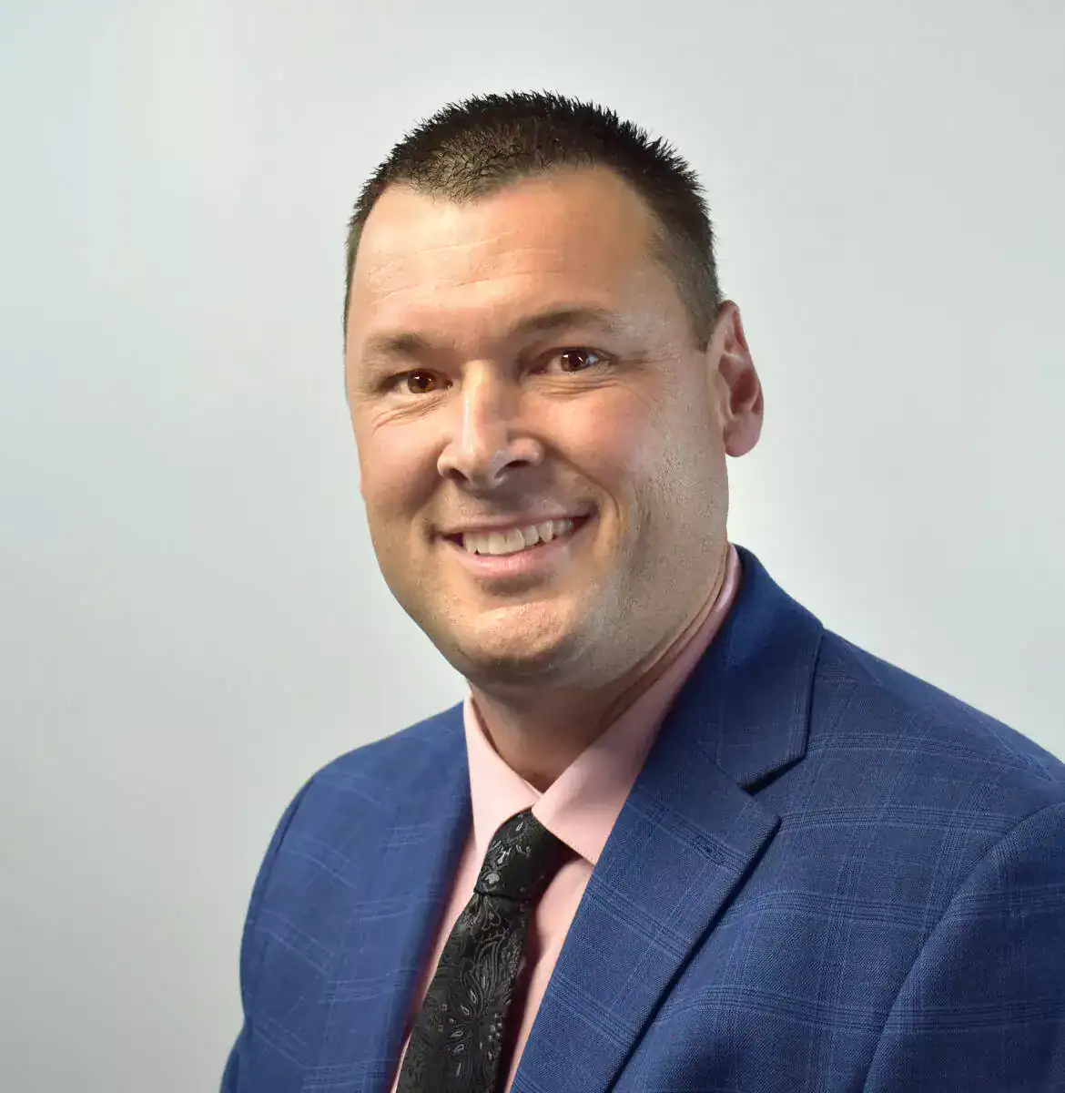 A smiling man with short dark hair, wearing a blue suit, pink shirt, and black patterned tie, posing against a plain light background.
