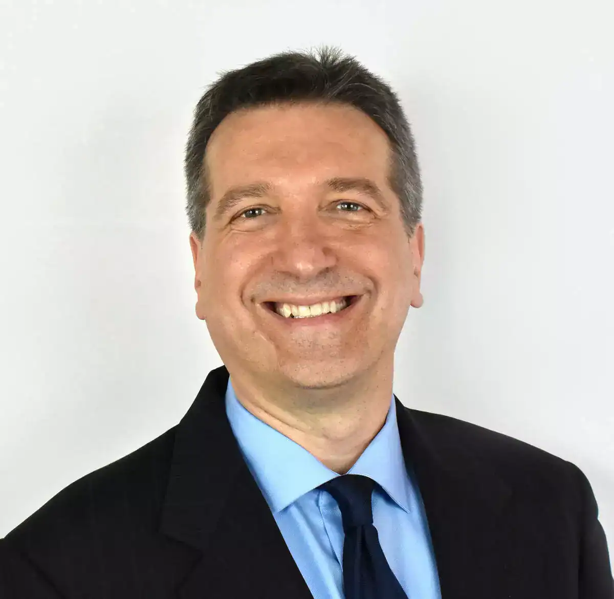 Smiling businessman in formal attire with a dark suit, light blue shirt, and dark tie, standing against a plain white background.