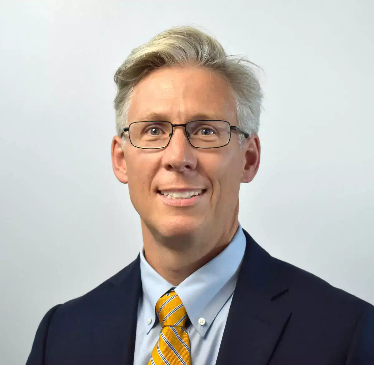 A professional businessman with glasses, wearing a navy suit, light blue shirt, and yellow striped tie, smiling at the camera.