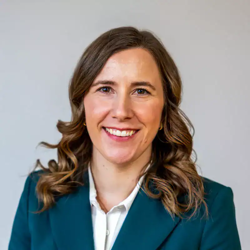 A smiling woman with wavy brown hair wearing a teal blazer and white shirt, looking directly at the camera against a plain light background.