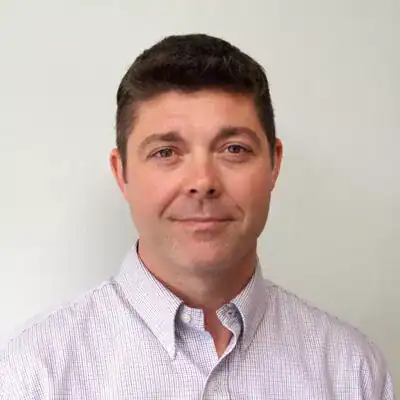 A professional headshot of a man with short dark hair, wearing a light-colored checkered shirt, smiling slightly against a plain background.