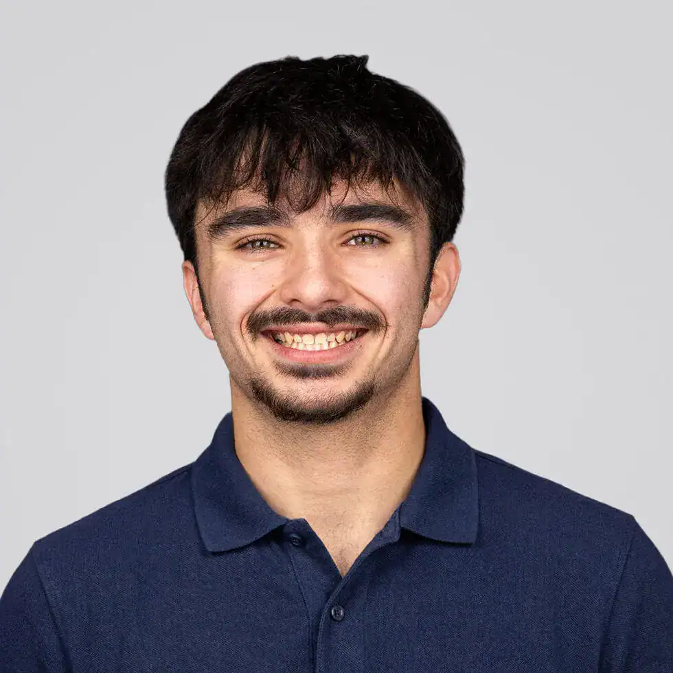 Portrait of a young man with dark hair and a beard, smiling and wearing a navy polo shirt against a light gray background.