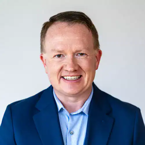 A smiling man in a blue suit and light blue shirt against a plain background.