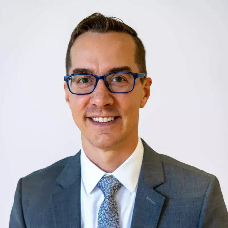 A professional man in a suit and glasses smiling at the camera against a plain white background.