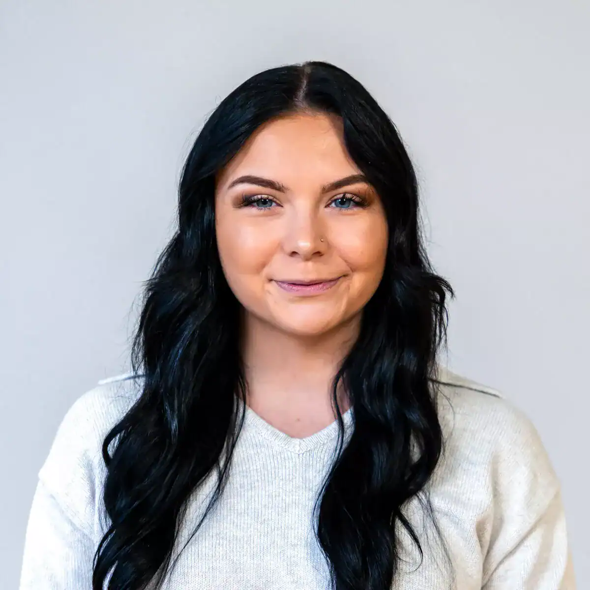 Close-up portrait of a young woman with long, wavy black hair, blue eyes, and a subtle smile, wearing a light-colored sweater against a plain background.