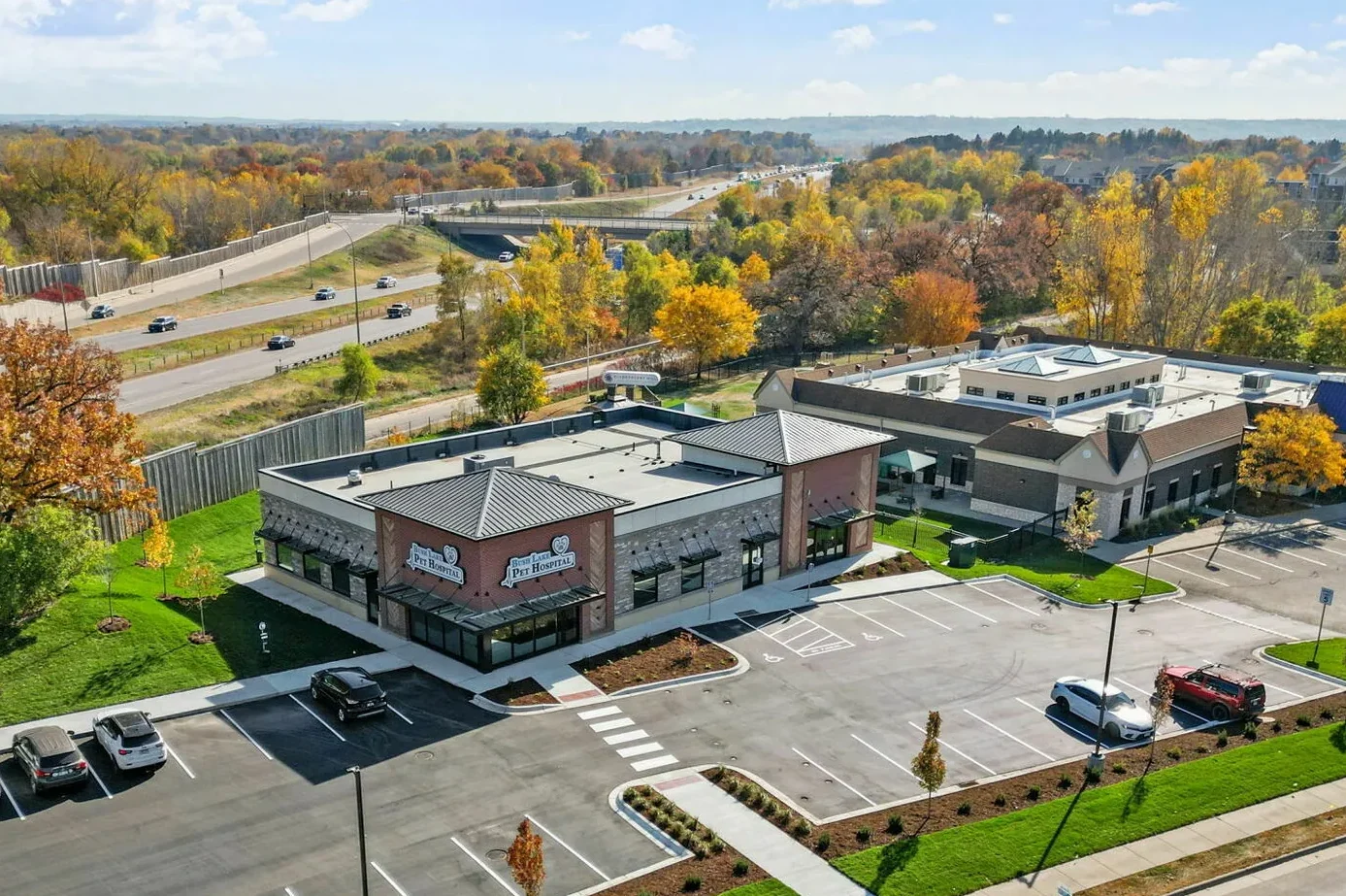 Aerial view of a retail shopping center with parking lot, surrounded by trees with fall foliage, and a highway in the background.