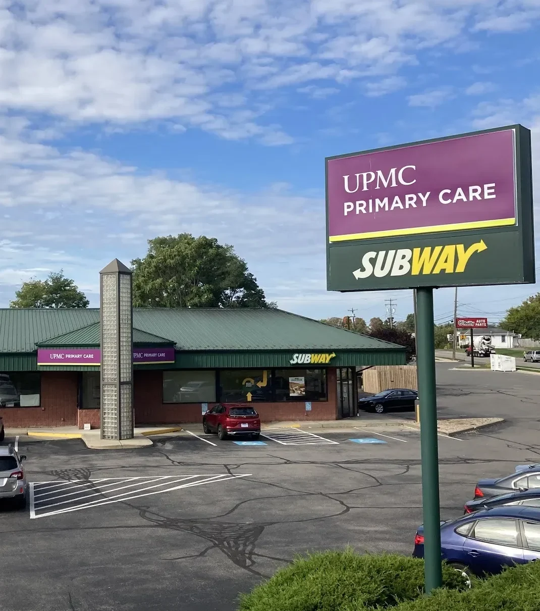 Exterior view of UPMC Primary Care and Subway clinic with parking lot and cars in front, under a partly cloudy sky.