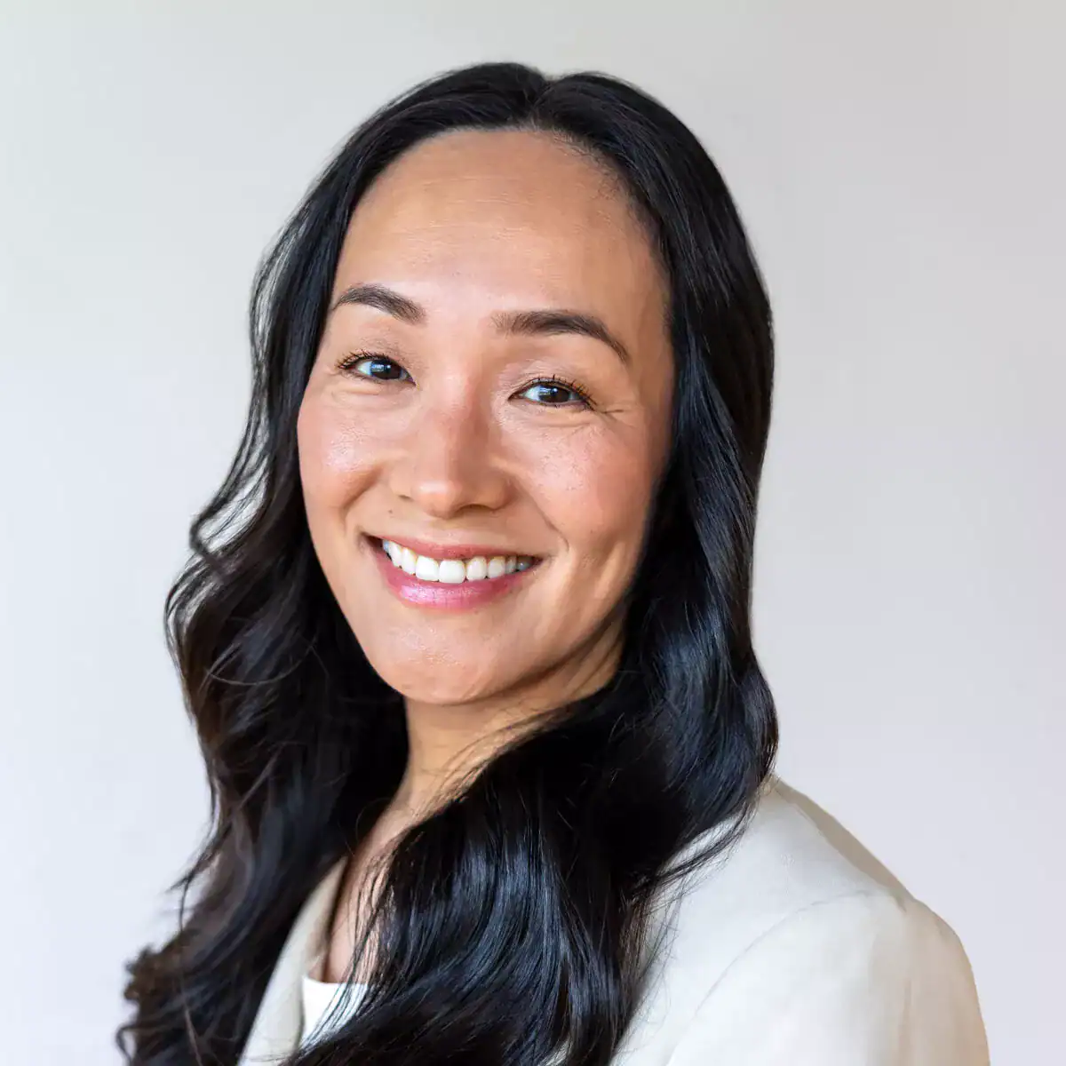 Close-up of a woman with long black hair, smiling and looking at the camera, wearing a light-colored blazer against a plain background.