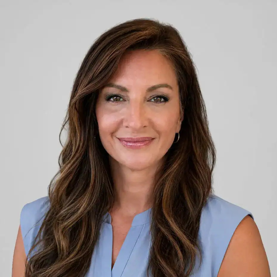 A professional woman with long wavy brown hair, wearing a light blue sleeveless blouse, smiling confidently at the camera against a plain light gray background.