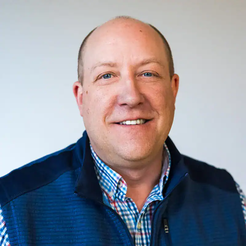 A smiling man with short hair and blue eyes, wearing a blue checkered shirt and a navy blue vest, looking directly at the camera.