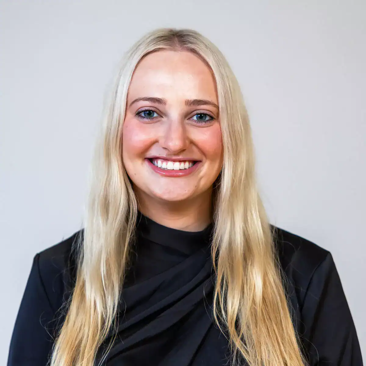 A young woman with long blonde hair, blue eyes, and a bright smile, wearing a black blazer against a plain light background.