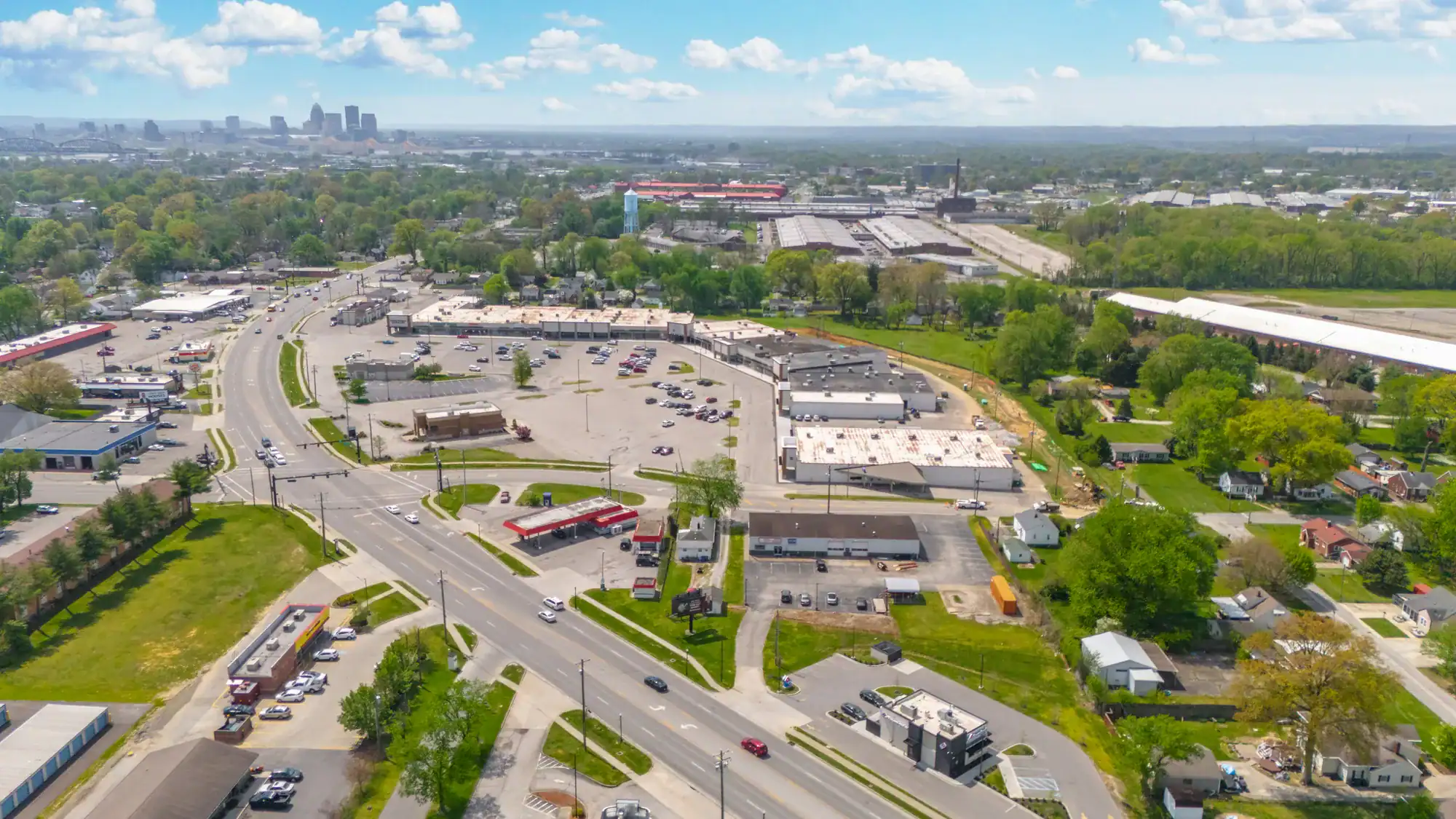 Aerial view of a shopping center with parking lot, roads, and surrounding greenery under a partly cloudy sky.