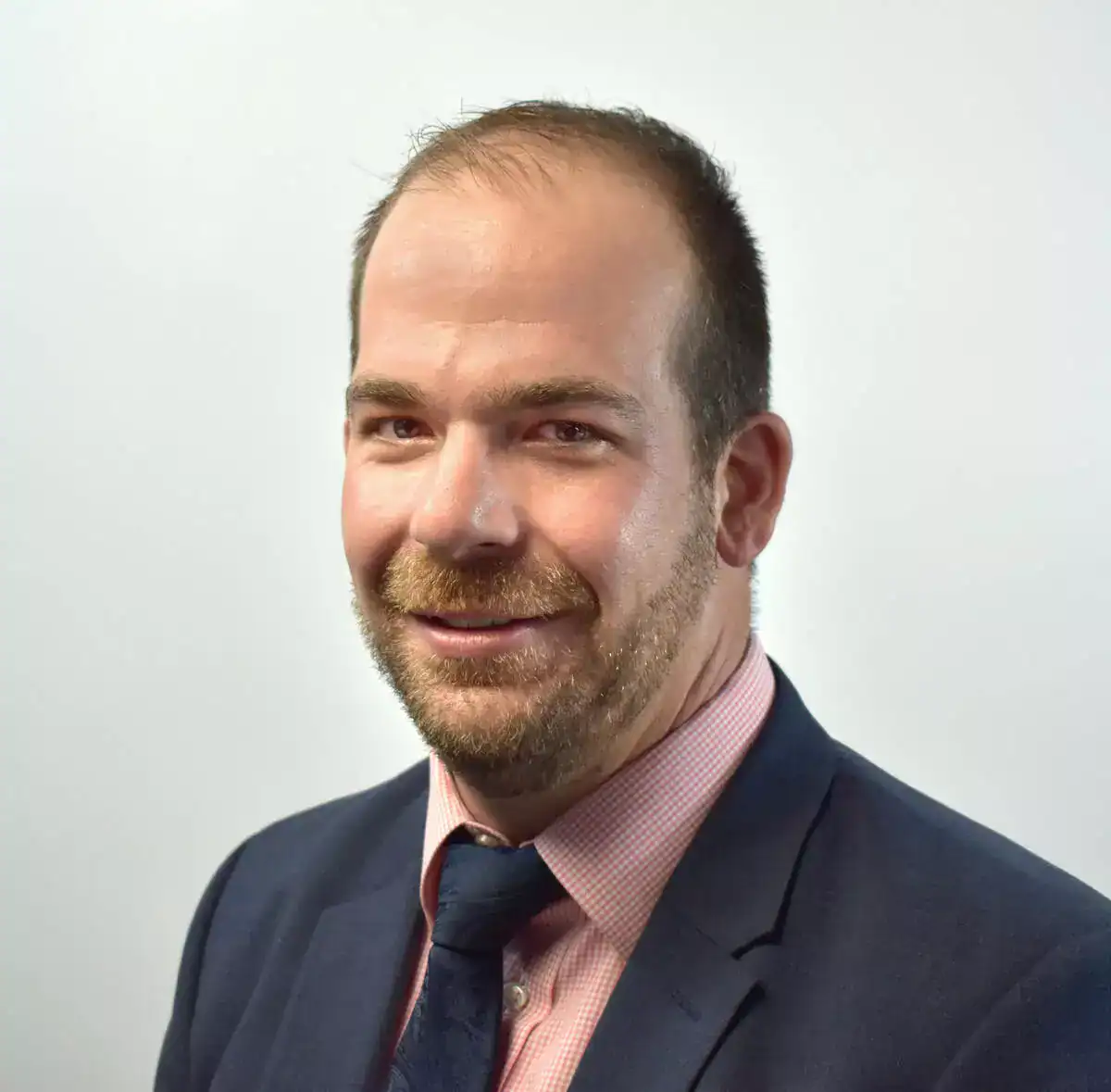 A professional man with a beard and short hair, wearing a navy suit, pink shirt, and dark tie, smiling at the camera against a plain white background.