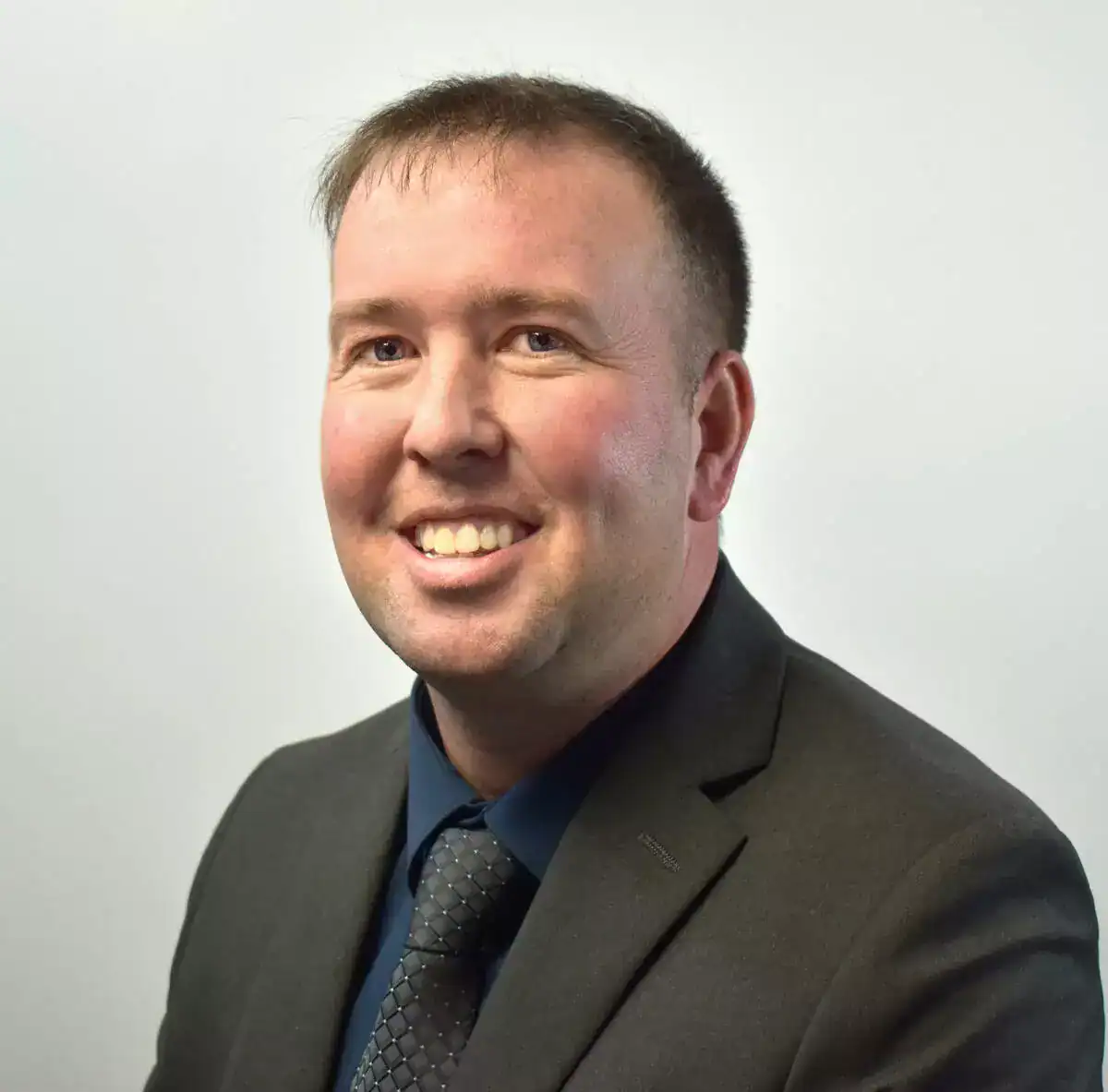 A smiling man in a dark suit, blue shirt, and patterned tie posing against a plain white background.
