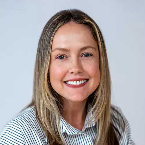 Close-up of a smiling woman with blonde hair and blue eyes, wearing a striped shirt against a plain background.