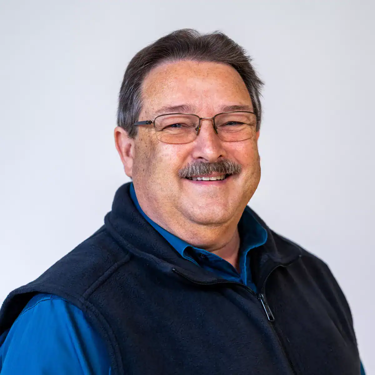 A middle-aged man with glasses, a mustache, and short dark hair, smiling at the camera against a plain white background.