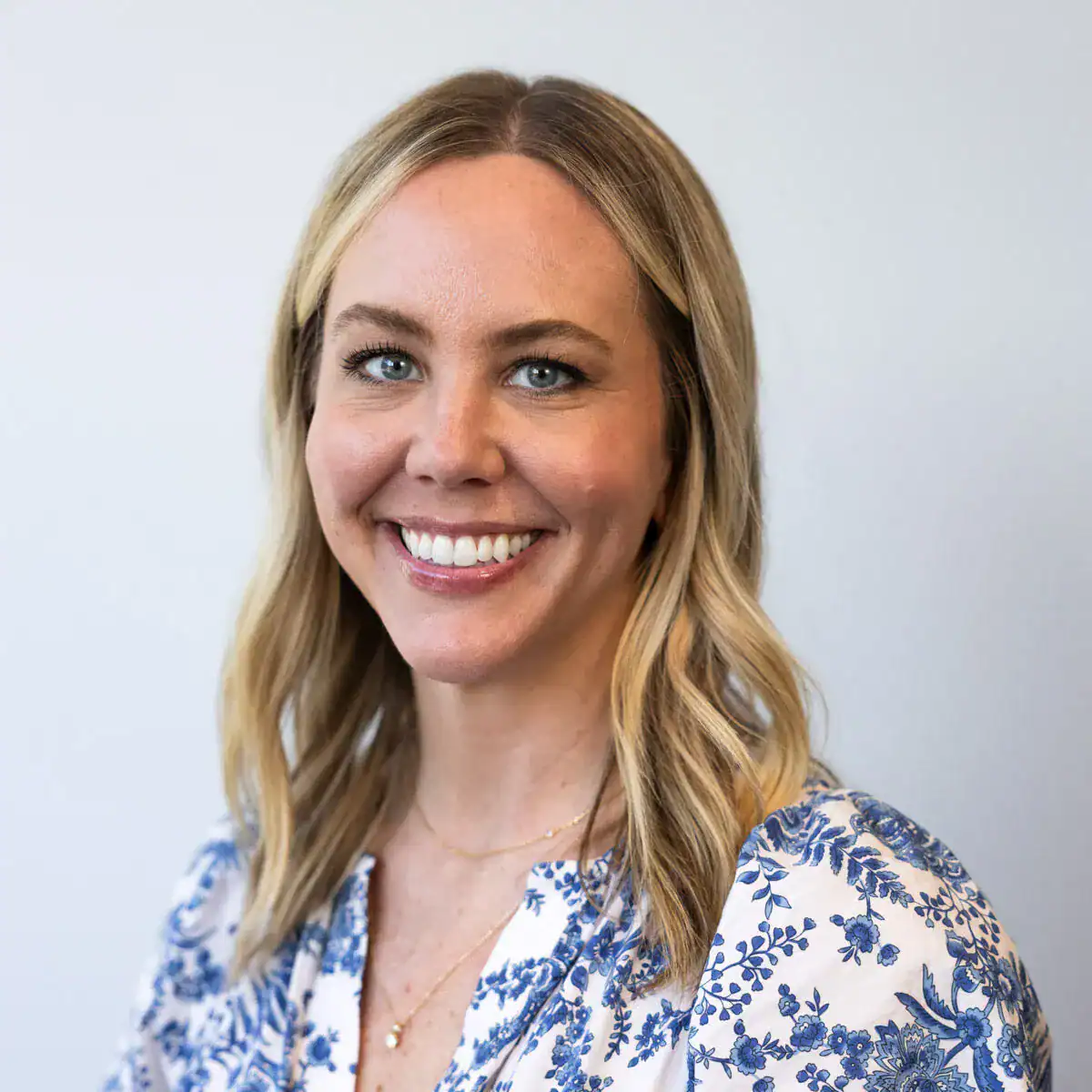 Portrait of a smiling woman with blonde hair, blue eyes, wearing a white and blue floral blouse, against a plain light background.