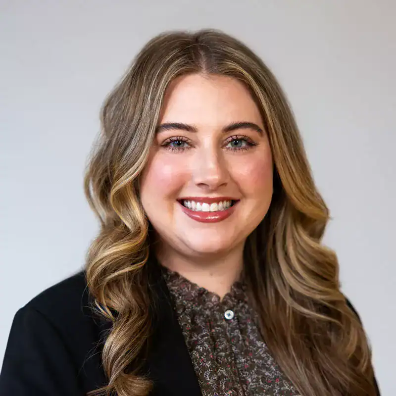 A young woman with long wavy blonde hair, smiling, wearing a black blazer and a patterned blouse against a neutral background.