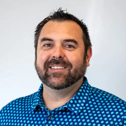 A professional headshot of a smiling man with dark hair and a beard, wearing a blue patterned shirt, against a plain light background.