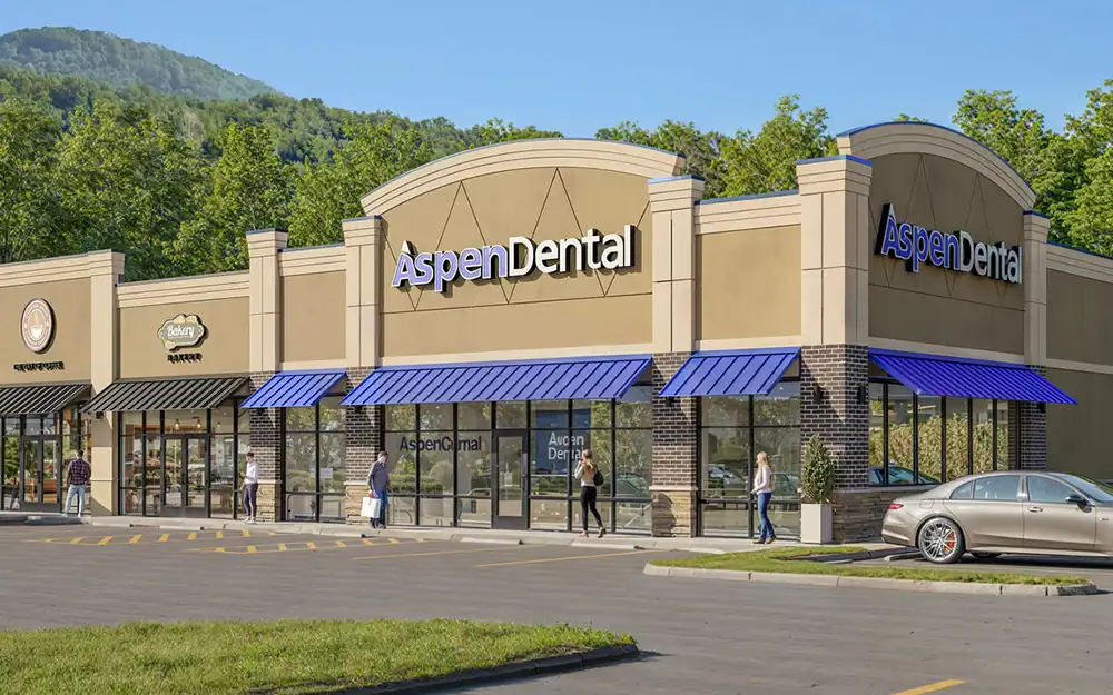 Exterior view of Aspen Dental clinic with storefronts, blue awnings, and people walking in front, set against a backdrop of green trees and a mountain.